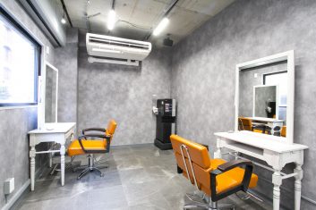 モダンなデザインの美容室内装、グレーの壁紙、オレンジの椅子、白いスタイリング台とミラーが特徴の広々としたサロン空間