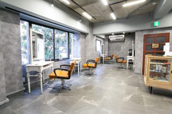 現代的なデザインの美容室の内部、洗練されたグレーテーマの壁とフロア、大きな窓と自然光、オレンジ色のアクセントが特徴的なスタイリングチェア