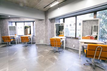 モダンな美容室の内装、明るい窓付きのスペースにオレンジ色の椅子と白いワークステーションが配置されたクリーンなデザイン