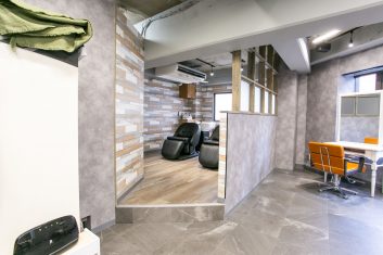 モダンな美容室の内装、洗髪用チェアと美容師の作業スペースが見え、ウッドとグレーを基調としたデザインが特徴