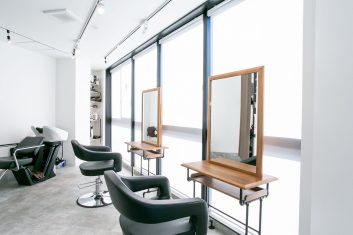 モダンで洗練された美容室の内装、自然光が入る大きな窓、シックなブラックのチェアと木製の鏡台が配置された快適な空間