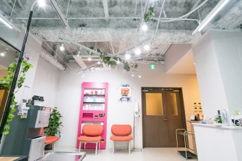 現代的な美容室の待合室、露出した天井と植物の装飾が特徴の明るく開放的な内装、赤い椅子とピンクの自販機装飾がアクセントになっている空間