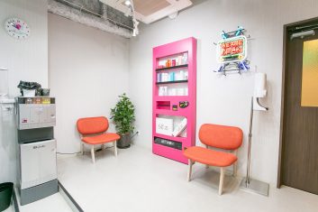 モダンで清潔な美容室の待合室、明るいピンクの製品展示棚とオレンジ色のチェアが特徴で、「WE ARE OPEN」のネオンサインが歓迎ムードを演出